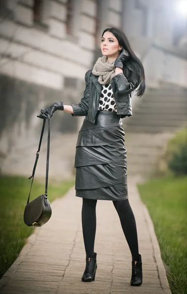 Attractive young woman in a winter fashion shot. Beautiful fashionable young girl in black leather waking on avenue. Elegant long hair brunette with handbag and scarf in urban scenery. — Stock Photo, Image