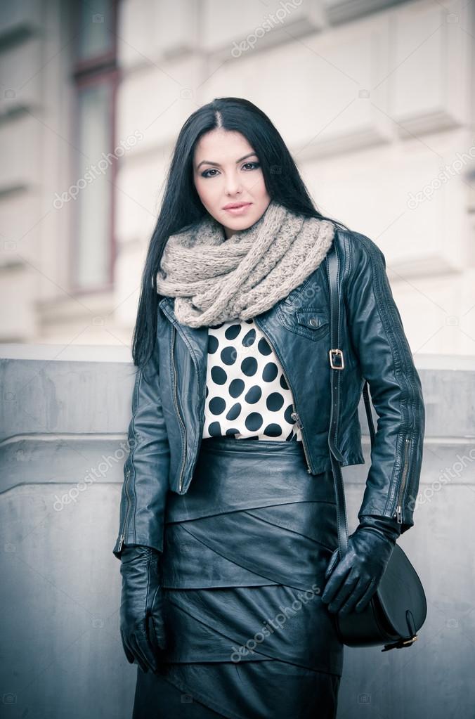 Attractive young woman in a winter fashion shot. Beautiful fashionable young girl in black leather on avenue. Elegant long hair brunette with handbag and scarf in urban scenery.
