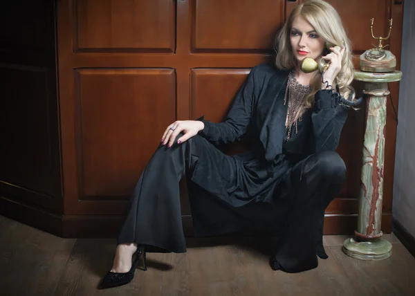 Mulher loira bonita nova em terno preto elegante falando por telefone sentado relaxado no chão. Menina cabelo claro sedutor inclinando-se no armário de madeira segurando um telefone vintage no interior clássico — Fotografia de Stock