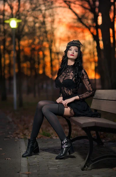 Schöne brünette Frau sitzt draußen im Park mit dramatischem Himmel im Hintergrund. modische Frau mit Mütze, die auf Bank ruht - Außenschuss. Vintage Lady in Schwarz posiert in Sonnenuntergangskulisse — Stockfoto