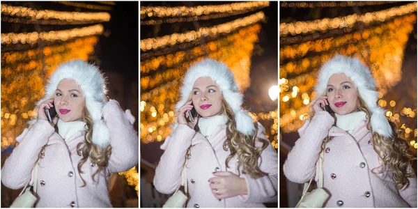 Portrait of young woman with white fur cap talking on mobile outdoor in cold winter evening. Beautiful blonde girl in winter clothes with Xmas lights in background. Cute female smiling, winter scenery — Zdjęcie stockowe
