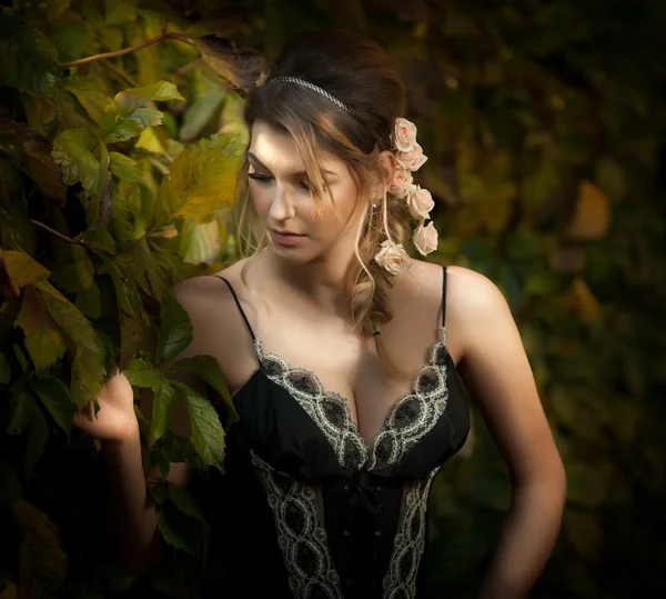 Hermosa mujer sensual con rosas en el pelo posando cerca de una pared de hojas verdes. Joven hembra en vestido elegante negro soñando despierto en la naturaleza. Atractiva dama voluptuosa con arreglo de pelo creativo —  Fotos de Stock