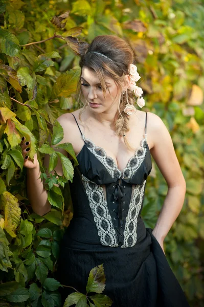 Mooie sensuele vrouw met rozen in haar poseren in de buurt van een muur van groene bladeren. Jonge vrouwelijke in elegante zwarte jurk dagdromen in de natuur. Aantrekkelijke wulpse dame met creatieve haar regeling — Stockfoto