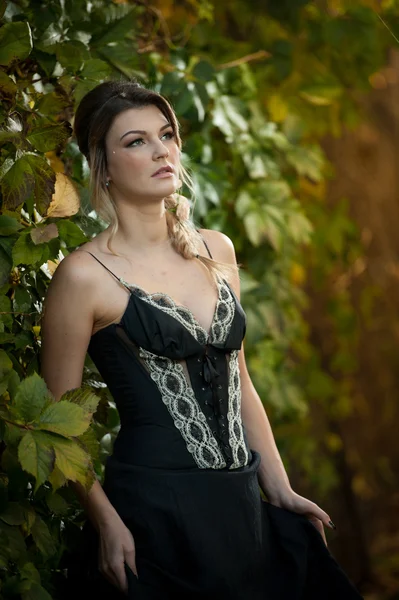 Beautiful sensual woman with roses in hair posing near a wall of green leaves. Young female in black elegant dress daydreaming in nature. Attractive voluptuous lady with creative hair arrangement — Stock Photo, Image