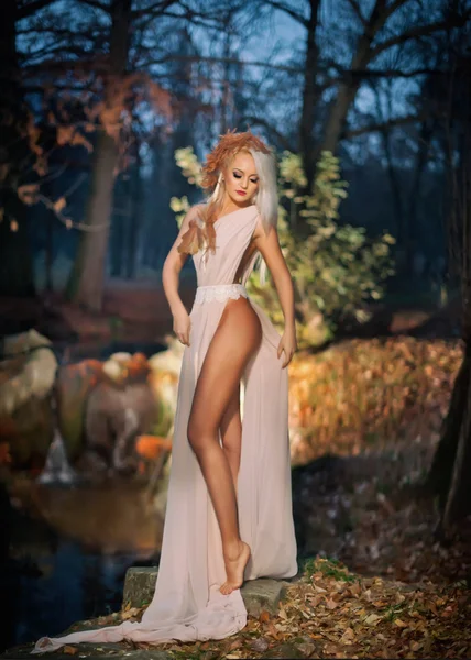 Encantadora joven de pie cerca del río en bosques encantados. Rubia sensual con vestido largo blanco posando provocativamente en bosque otoñal. Chica con aspecto de hada en el paisaje de otoño. Mujer romántica cerca del lago — Foto de Stock