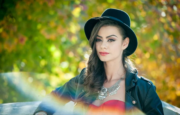 Hermosa mujer con sombrero negro posando en el parque otoñal. Joven morena pasando tiempo durante el otoño en el bosque. Chica atractiva de pelo largo con maquillaje creativo y vestido rojo, al aire libre tiro durante el otoño — Foto de Stock