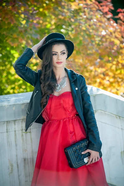 Beautiful woman with black hat posing in autumnal park. Young brunette spending time during autumn in forest. Long hair attractive girl with creative makeup and red dress, outdoors shot during fall — Stock Photo, Image