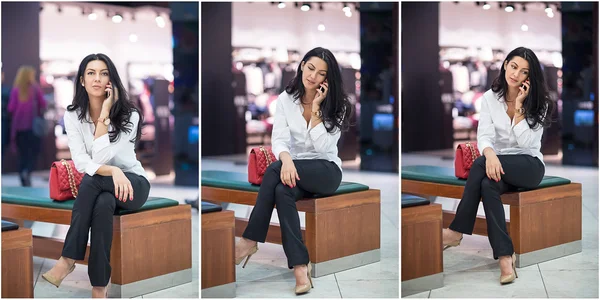 Attractive woman speaking on mobile in mall. Beautiful fashionable young girl in white male shirt sitting on wooden bench in modern shopping center. Long hair brunette posing smiling talking by phone. — Stock Photo, Image