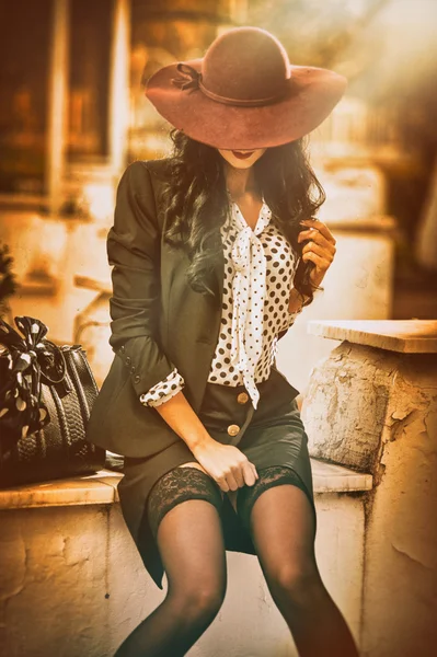 Attraente giovane donna con cappello grande di colore bordeaux in modo autunnale girato. Bella signora in abito nero con gonna corta e calze sexy seduto sulla parete di marmo. Elegante bruna all'aperto . — Foto Stock