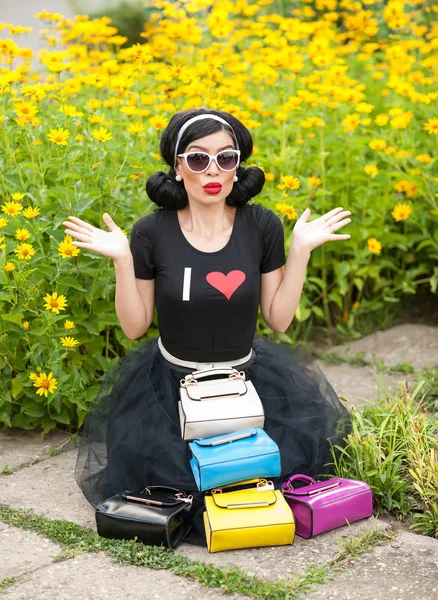 Hermosa chica con aspecto retro con un traje negro que se divierte en el parque mostrando una gran cantidad de bolsas de colores. Morena de moda con gafas de sol y bolsos delante, flores amarillas en el fondo — Foto de Stock