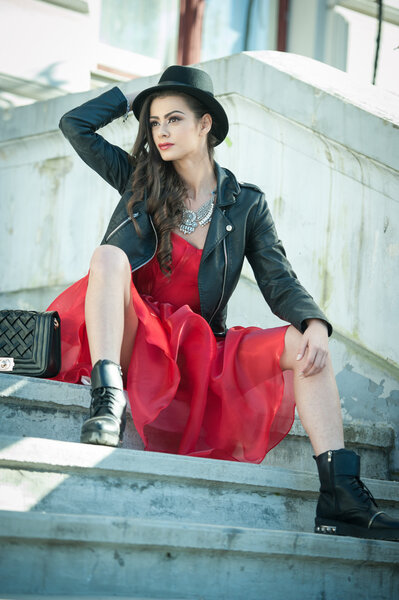 Beautiful woman with black hat, red dress and boots posing sitting on stairs. Young brunette spending time during autumn. Long hair attractive girl with creative makeup and red dress, outdoors shot