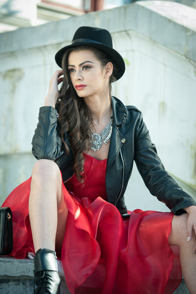 Beautiful woman with black hat, red dress and boots posing sitting on stairs. Young brunette spending time during autumn. Long hair attractive girl with creative makeup and red dress, outdoors shot