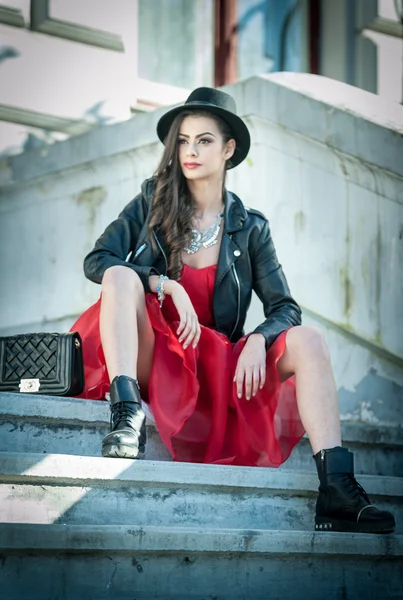 Mulher bonita com chapéu preto, vestido vermelho e botas posando sentado nas escadas. Jovem morena passando tempo durante o outono. Cabelo longo menina atraente com maquiagem criativa e vestido vermelho, tiro ao ar livre — Fotografia de Stock