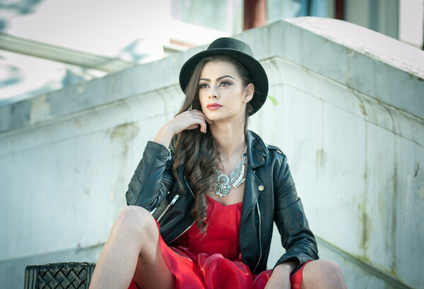 Beautiful woman with black hat, red dress and boots posing sitting on stairs. Young brunette spending time during autumn. Long hair attractive girl with creative makeup and red dress, outdoors shot