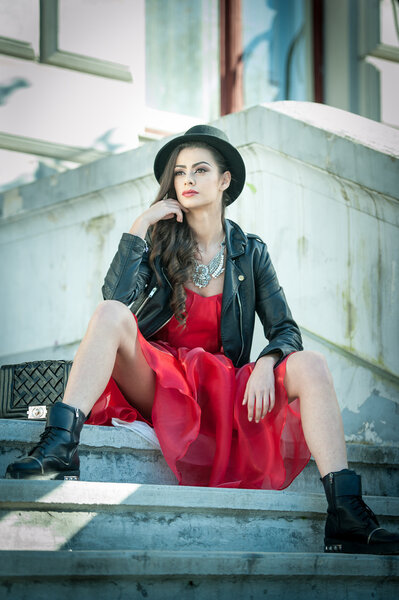 Женщина в черной шляпе, красном платье и сапогах позирует сидя на лестнице. Осенью юная брюнетка проводит время. Длинные волосы привлекательная девушка с творческим макияжем и красным платьем, на открытом воздухе выстрел
