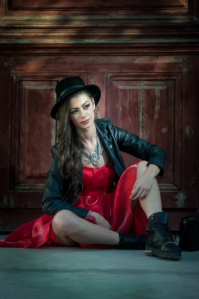 Jonge mooie brunette vrouw met rode korte jurk en zwarte hoed poseren sensuele in vintage landschap. Romantische mysterieuze jonge dame ontspannen zittend op de vloer tegen vintage houten muur — Stockfoto