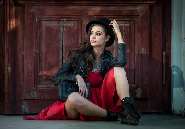 Joven mujer morena hermosa con vestido corto rojo y sombrero negro posando sensual en el paisaje vintage. Romántico misteriosa joven relajante sentado en el suelo contra la pared de madera vintage — Foto de Stock