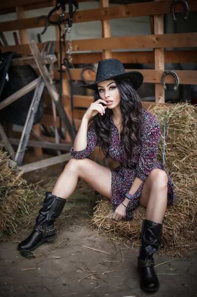 Schöne Brünette mit Country-Look, drinnen in stabilem, rustikalem Stil gedreht. attraktive Frau mit schwarzem Cowboyhut, kurzem Kleid und Stiefeln, Bauer im amerikanischen Landhausstil mit Stallgeschirr im Hintergrund — Stockfoto