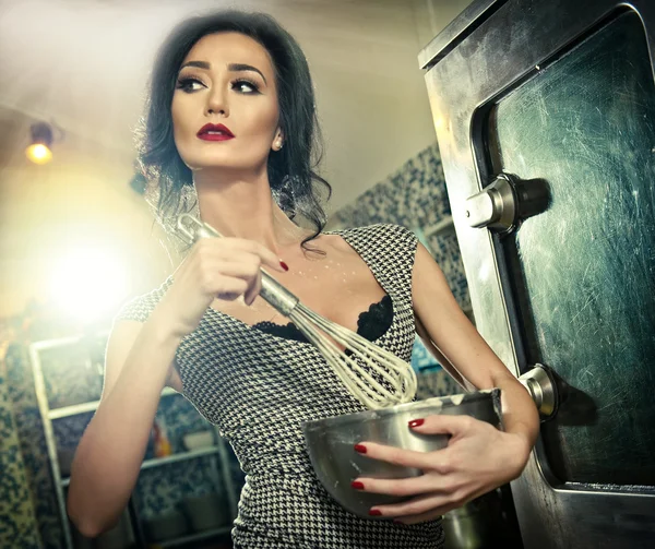 Beautiful brunette mixing ingredients in a bowl. Sensual slim young woman with black bra and low cut neck baking in a professional kitchen. Attractive lady with makeup cooking holding a whisk — Stockfoto