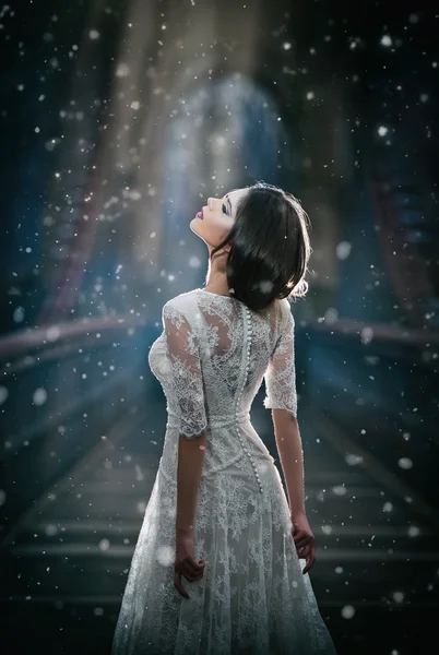Bella giovane signora indossa elegante abito bianco godendo le travi di luce celeste e fiocchi di neve che cadono sul suo viso. Bella ragazza bruna in abito da sposa lungo in posa su un ponte in paesaggio invernale — Foto Stock