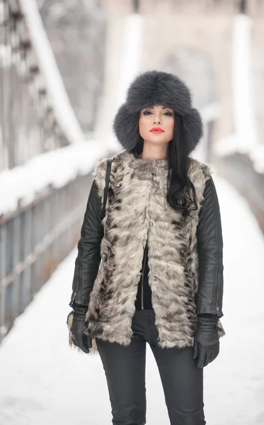 Mujer atractiva con gorra de piel negra y chaleco gris disfrutando del invierno. Vista frontal de la chica morena de moda posando en un puente cubierto de nieve. Hermosa joven hembra con traje de clima frío —  Fotos de Stock