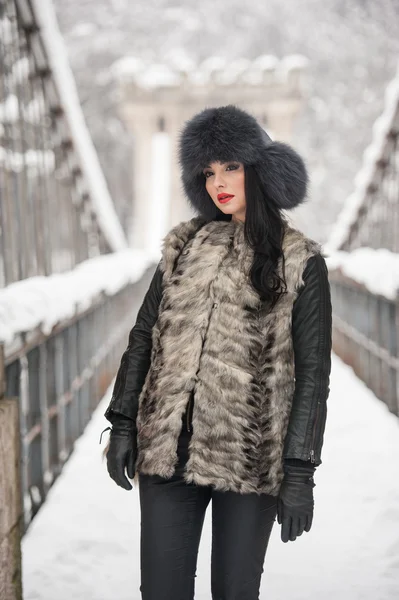 Attractive woman with black fur cap and gray waistcoat enjoying the winter. Frontal view of fashionable brunette girl posing on a snow covered bridge. Beautiful young female with cold weather outfit — Zdjęcie stockowe