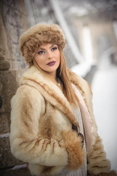 Aantrekkelijke vrouw met bruin bont GLB en genieten van de winter jas. Zijaanzicht van modieus blond meisje poseren tegen sneeuw overdekte brug. Mooie jonge vrouw met koud weer outfit — Stockfoto