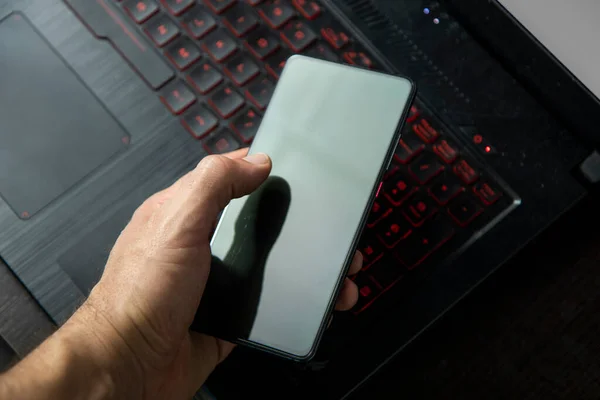 hand of a man using a black cell phone and background a black laptop with red lights