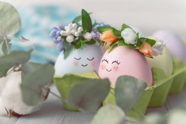 Paskalya konsepti. Pastel renklerle boyanmış Paskalya yumurtaları. Üzerinde çelenkler olan yumurta tepsisinde sevimli yüzler. Hafif arkaplan, metin için yer.