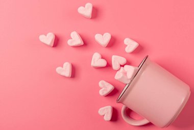 Pembe bir kupa yan yatar pembe bir zemin üzerinde, açık pembe marşmelovlar serpiştirilmiş kalpler şeklinde