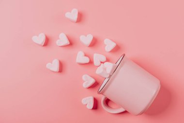 Pembe bir kupa yan yatar pembe bir zemin üzerinde, açık pembe marşmelovlar serpiştirilmiş kalpler şeklinde