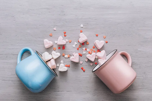 two mugs of blue and pink lie on their side, small candy hearts and marshmallow hearts spilled out