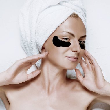 Gözlerinin altındaki deri için kozmetik maske. Başında havlu olan bir kıza göz maskesi tak.