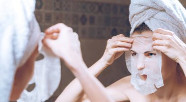 Kadın, kafasında havluyla aynanın yanında durur ve kozmetik bir maske takar.