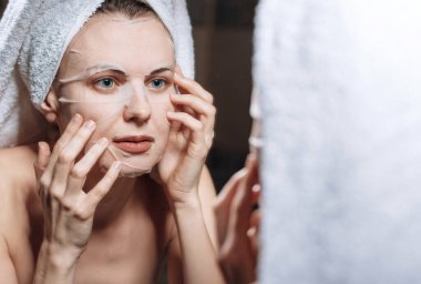 Kadın, kafasında havluyla aynanın yanında durur ve kozmetik bir maske takar.