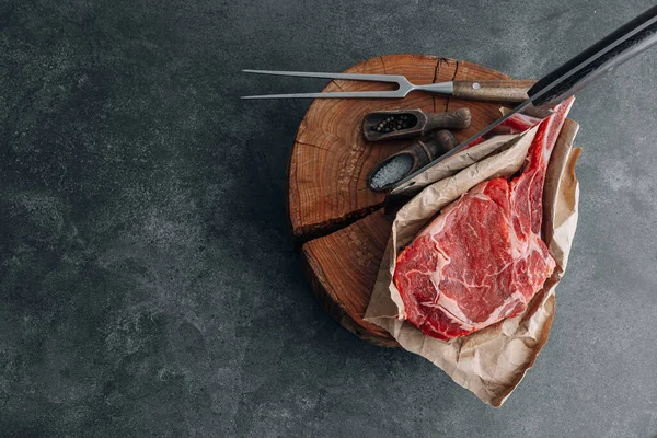 on a wooden block for meat a fresh raw tamahawk steak or a cowboy steak with a butcher\'s chopping ax for meat, next to it is a mixture of peppers and coarse salt, milled with thyme. fresh beef steak