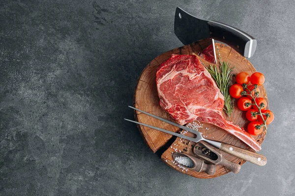 on a wooden block for meat a fresh raw tamahawk steak or a cowboy steak with a butcher\'s chopping ax for meat, next to it is a mixture of peppers and coarse salt, milled with thyme. fresh beef steak