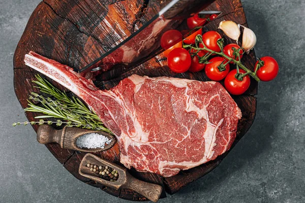 on a wooden block for meat a fresh raw tamahawk steak or a cowboy steak with a butcher\'s chopping ax for meat, next to it is a mixture of peppers and coarse salt, milled with thyme. fresh beef steak