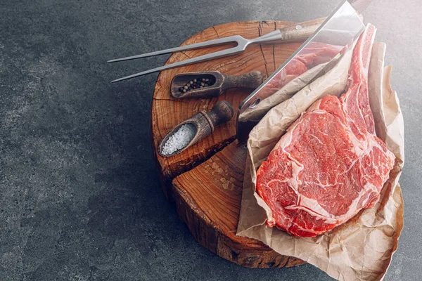 on a wooden block for meat a fresh raw tamahawk steak or a cowboy steak with a butcher\'s chopping ax for meat, next to it is a mixture of peppers and coarse salt, milled with thyme. fresh beef steak