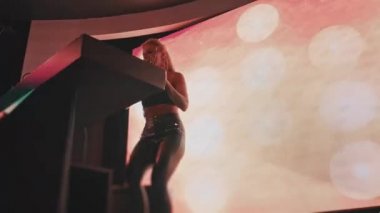 Avrupalı havalı bir bayan DJ, sarı saçlı ve Hıristiyan haçlarından yapılmış şık bir maske, bir gece kulübünde çalışıyor ve dans müziği yaratıyor..