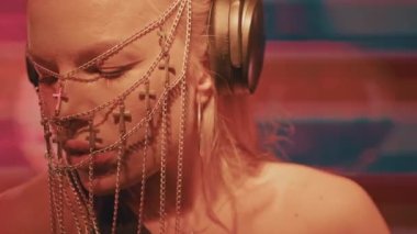 Avrupalı havalı bir bayan DJ, sarı saçlı ve Hıristiyan haçlarından yapılmış şık bir maske, bir gece kulübünde çalışıyor ve dans müziği yaratıyor..