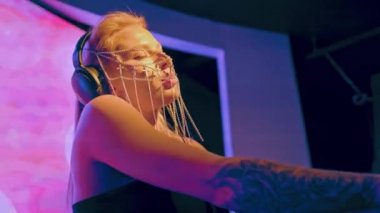Avrupalı havalı bir bayan DJ, sarı saçlı ve Hıristiyan haçlarından yapılmış şık bir maske, bir gece kulübünde çalışıyor ve dans müziği yaratıyor..