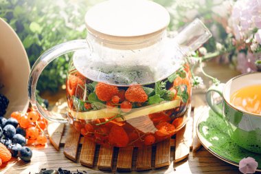 Şeffaf bir çaydanlıkta doğal aromatik meyve çayı ve böğürtlen ve çiçeklerin arasında bir fincanda. Böğürtlen ve kır çiçekleri aromalı sıcak aromatik çay..