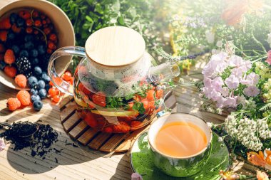 Şeffaf bir çaydanlıkta doğal aromatik meyve çayı ve böğürtlen ve çiçeklerin arasında bir fincanda. Böğürtlen ve kır çiçekleri aromalı sıcak aromatik çay..