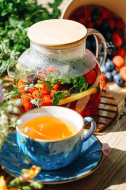 Şeffaf bir çaydanlıkta doğal aromatik meyve çayı ve böğürtlen ve çiçeklerin arasında bir fincanda. Böğürtlen ve kır çiçekleri aromalı sıcak aromatik çay..