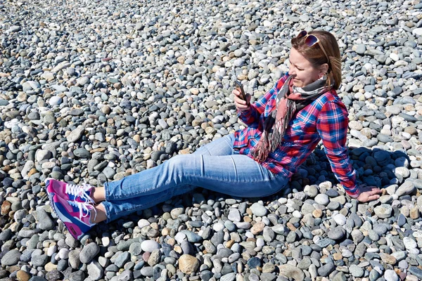 Hermosa chica con teléfono sentado en la playa rocosa — Foto de Stock