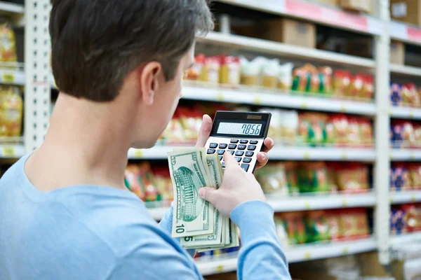 Hombre con dólares y calculadora considera los costos en la tienda — Foto de Stock