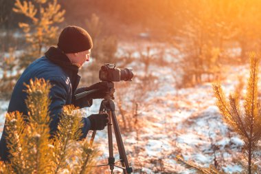 Kışın kalın giyinmiş bir adam tripodunda dijital kamerayla güzel çekimler yapar. Gün batımında bir portre, kış fotoğrafları.