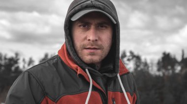 Şapkalı ve kapüşonlu bir adamın portresi. Yüzünde kıllar var. Ormanın arka planında ciddi bir yüzü olan bir adam.