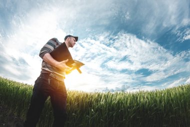 Genç bir tarım uzmanı yeşil buğday tarlasında elinde bir klasör tutuyor. Bir çiftçi gün batımında tarımın arka planına not alır. Belge klasörlü şapkalı bir adam.